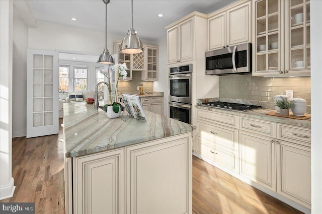 kitchen featuring decorative light fixtures, light stone counters, stainless steel appliances, a center island with sink, and cream cabinetry