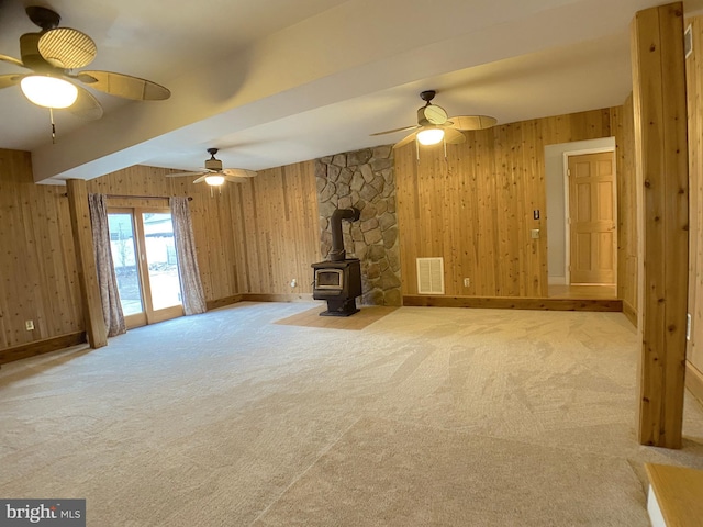 unfurnished living room with ceiling fan, beam ceiling, wooden walls, carpet floors, and a wood stove