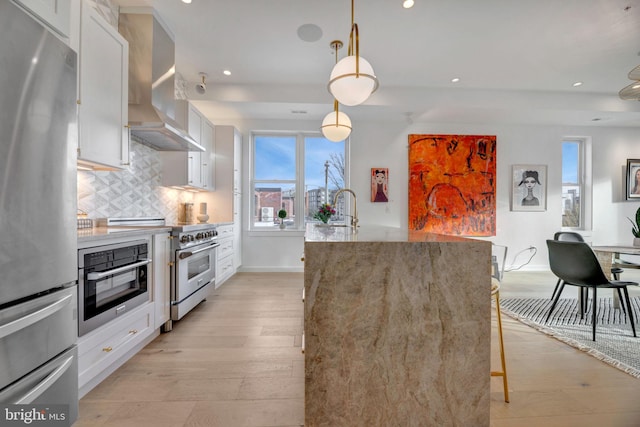 kitchen with appliances with stainless steel finishes, decorative light fixtures, wall chimney range hood, and white cabinets