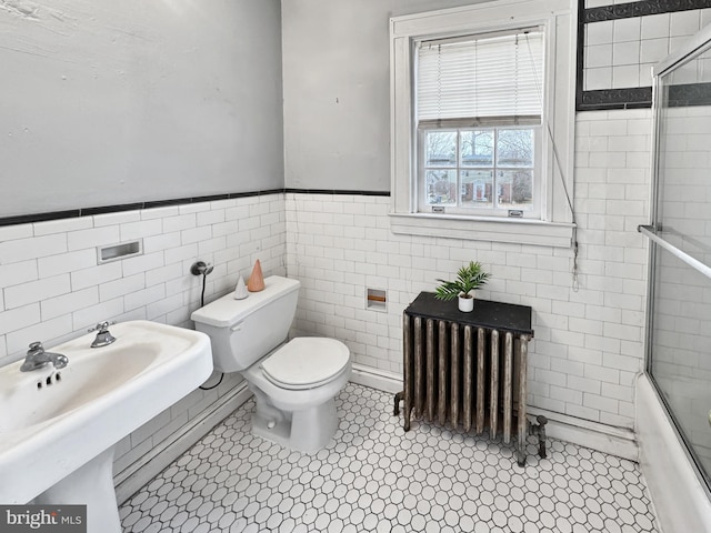 full bathroom featuring combined bath / shower with glass door, radiator heating unit, sink, tile walls, and toilet