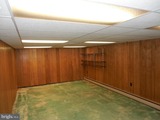 basement featuring a paneled ceiling, wooden walls, and a baseboard heating unit