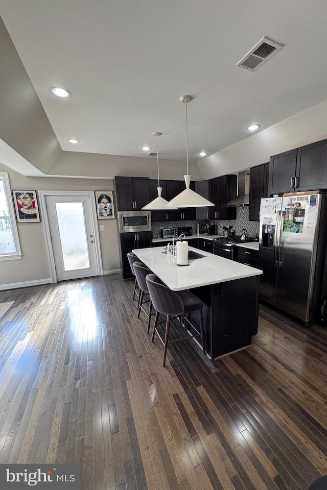 kitchen with wall chimney range hood, sink, hanging light fixtures, stainless steel appliances, and a center island with sink