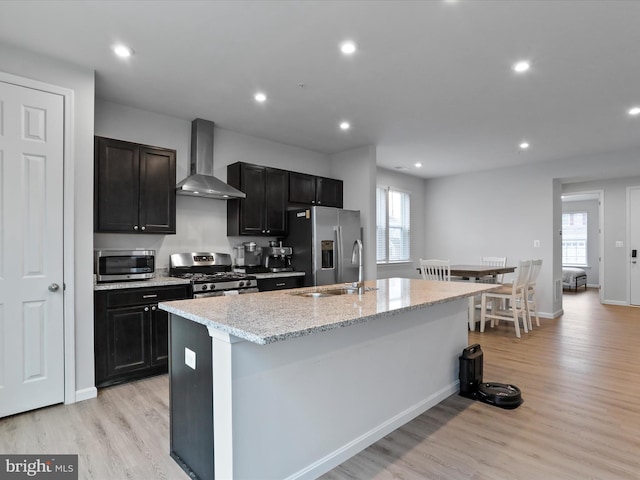 kitchen with wall chimney exhaust hood, stainless steel appliances, light hardwood / wood-style floors, and a kitchen island with sink