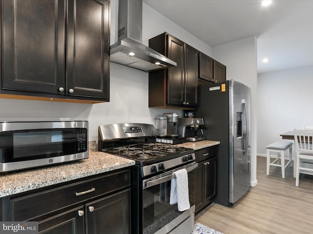 kitchen with appliances with stainless steel finishes, dark brown cabinetry, light stone counters, light wood-type flooring, and wall chimney exhaust hood