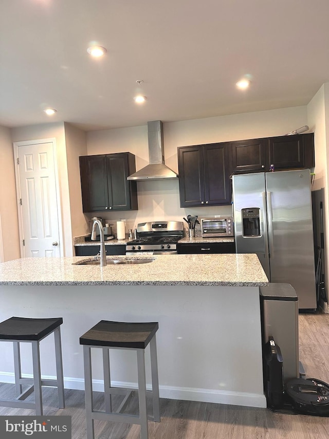 kitchen with hardwood / wood-style flooring, stainless steel appliances, light stone countertops, a center island with sink, and wall chimney exhaust hood