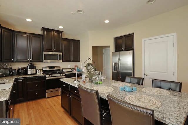 kitchen with appliances with stainless steel finishes, an island with sink, a kitchen bar, light stone countertops, and light wood-type flooring