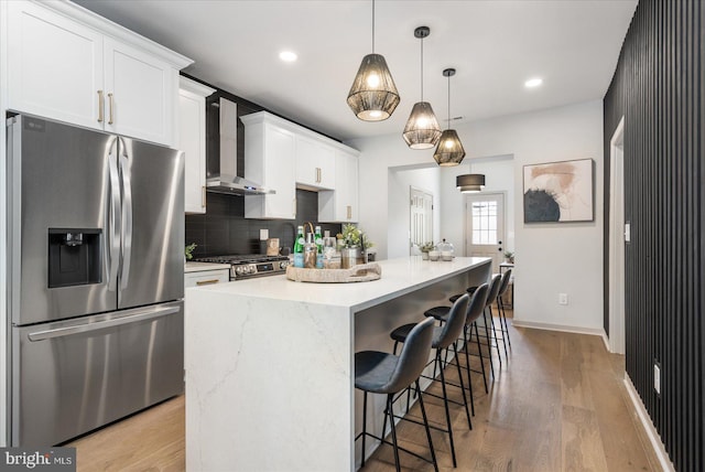 kitchen with wall chimney exhaust hood, white cabinetry, an island with sink, pendant lighting, and stainless steel appliances