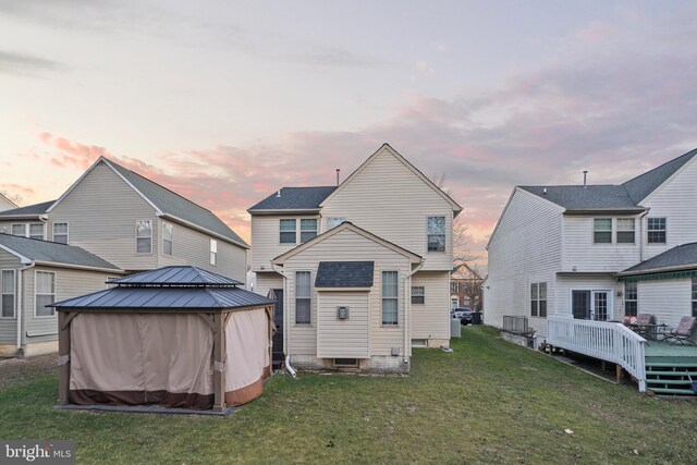 back house at dusk featuring a wooden deck, a gazebo, and a lawn