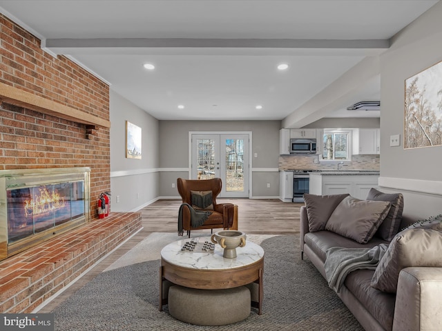 living room featuring french doors, sink, a brick fireplace, light hardwood / wood-style flooring, and beamed ceiling