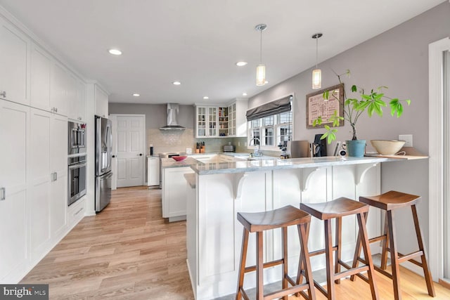 kitchen featuring white cabinetry, hanging light fixtures, stainless steel appliances, kitchen peninsula, and wall chimney exhaust hood