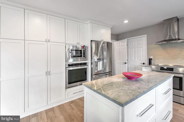 kitchen with wall chimney range hood, stainless steel appliances, light hardwood / wood-style floors, white cabinets, and a kitchen island