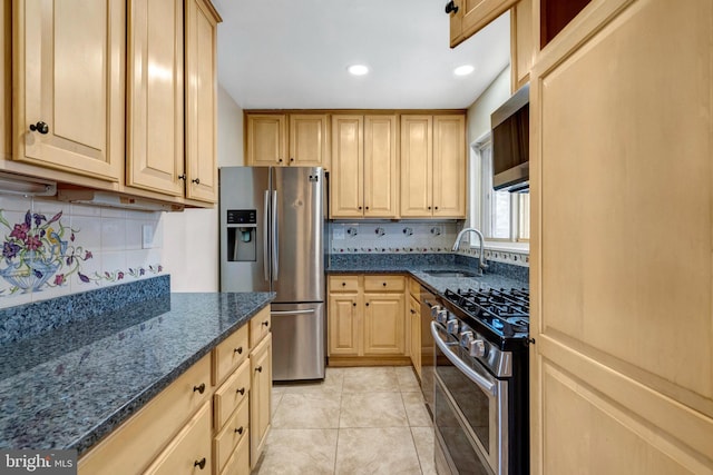 kitchen with light tile patterned floors, dark stone countertops, stainless steel appliances, light brown cabinetry, and a sink