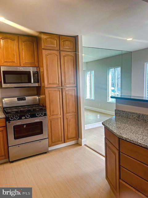 kitchen featuring light wood finished floors, baseboards, brown cabinetry, appliances with stainless steel finishes, and dark stone countertops