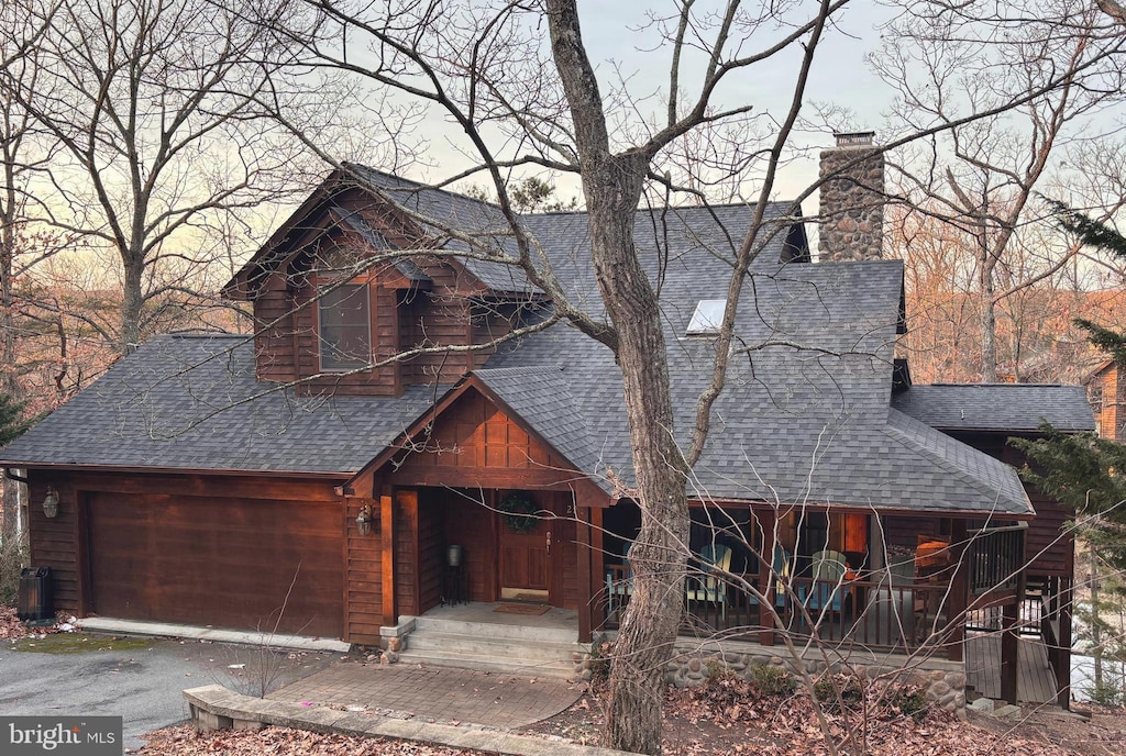 chalet / cabin with a chimney, aphalt driveway, roof with shingles, an attached garage, and a porch