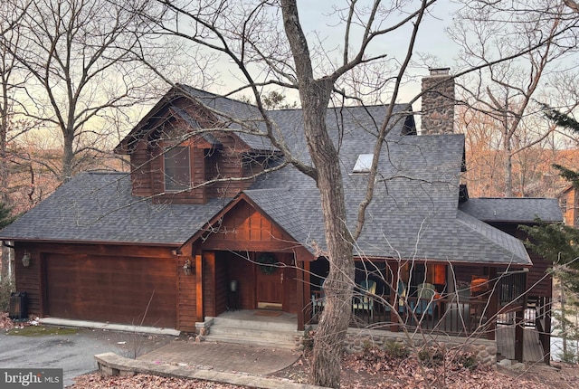 chalet / cabin with a chimney, aphalt driveway, roof with shingles, an attached garage, and a porch