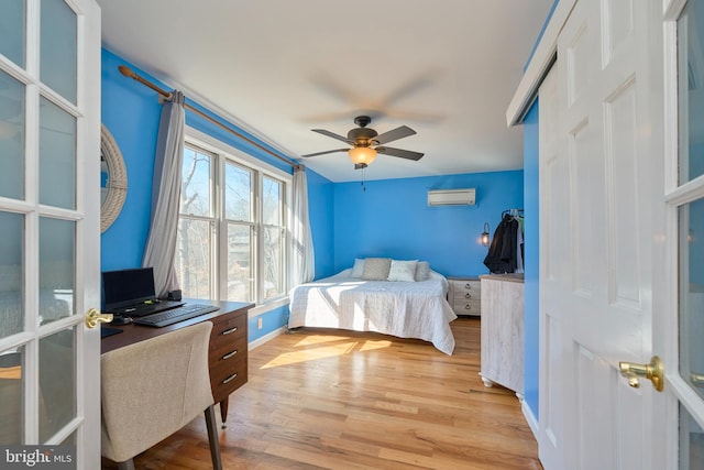 bedroom featuring light wood finished floors, ceiling fan, baseboards, and a wall mounted air conditioner