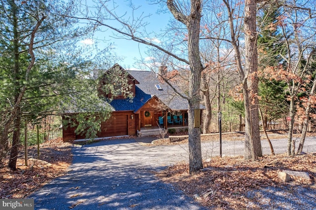 rustic home featuring a porch, an attached garage, driveway, roof with shingles, and a chimney