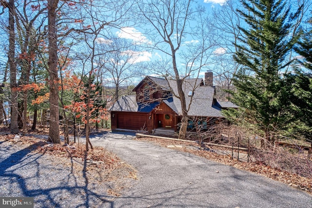 chalet / cabin with an attached garage, driveway, a chimney, and roof with shingles