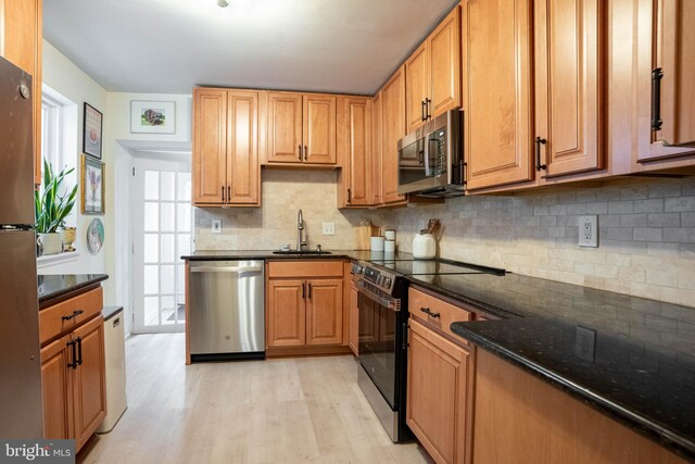 kitchen with sink, light hardwood / wood-style flooring, dark stone countertops, appliances with stainless steel finishes, and backsplash