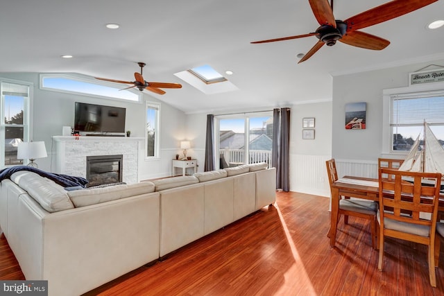 living room featuring a stone fireplace, crown molding, hardwood / wood-style flooring, ceiling fan, and vaulted ceiling with skylight