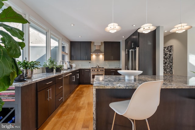 kitchen featuring hanging light fixtures, dark brown cabinets, high quality fridge, oven, and wall chimney exhaust hood