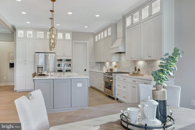 kitchen with premium appliances, an island with sink, custom range hood, white cabinets, and decorative light fixtures