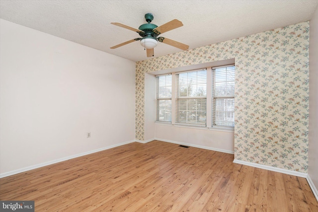 spare room with a textured ceiling, ceiling fan, baseboards, light wood finished floors, and wallpapered walls