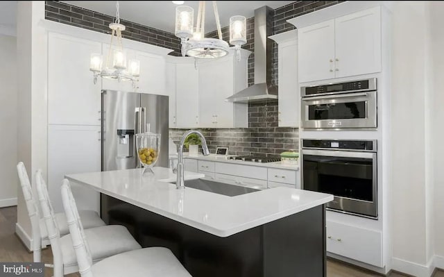 kitchen with wall chimney range hood, appliances with stainless steel finishes, white cabinetry, hanging light fixtures, and an island with sink