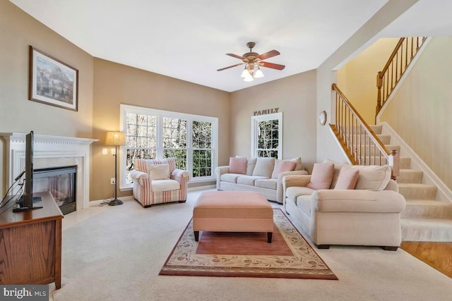 living area with baseboards, light colored carpet, a glass covered fireplace, ceiling fan, and stairs