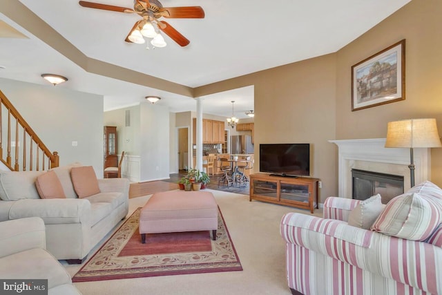 living area featuring stairs, ceiling fan with notable chandelier, light colored carpet, and a glass covered fireplace