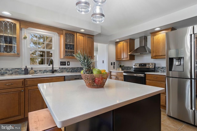 kitchen with a sink, wall chimney range hood, appliances with stainless steel finishes, brown cabinets, and glass insert cabinets