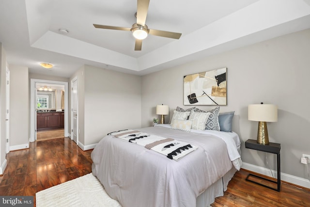 bedroom with dark wood-type flooring, ceiling fan, a tray ceiling, and ensuite bath