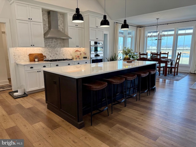 kitchen featuring wall chimney range hood, stainless steel appliances, a spacious island, white cabinets, and light wood-type flooring