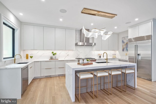 kitchen with wall chimney exhaust hood, sink, white cabinetry, a center island with sink, and appliances with stainless steel finishes