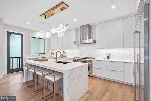 kitchen featuring an island with sink, sink, white cabinets, high end appliances, and wall chimney exhaust hood