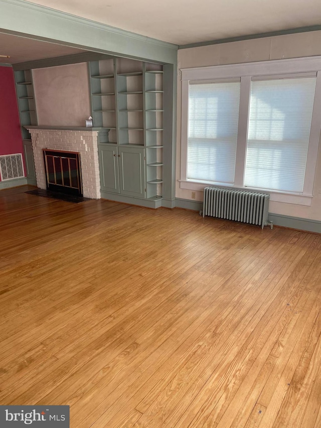 unfurnished living room with crown molding, light hardwood / wood-style flooring, radiator heating unit, a brick fireplace, and built in shelves