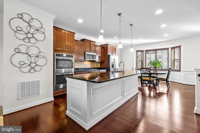 kitchen featuring a center island with sink, stainless steel appliances, visible vents, hanging light fixtures, and brown cabinetry