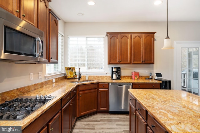 kitchen featuring a sink, stainless steel appliances, light wood finished floors, light stone countertops, and hanging light fixtures