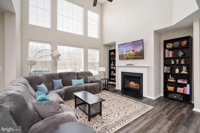 living room featuring baseboards, dark wood finished floors, a fireplace with flush hearth, built in features, and a ceiling fan