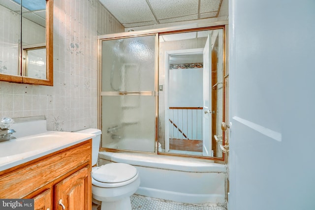 full bathroom with toilet, a paneled ceiling, bath / shower combo with glass door, tile walls, and vanity