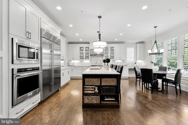 kitchen with pendant lighting, white cabinetry, sink, built in appliances, and a center island with sink