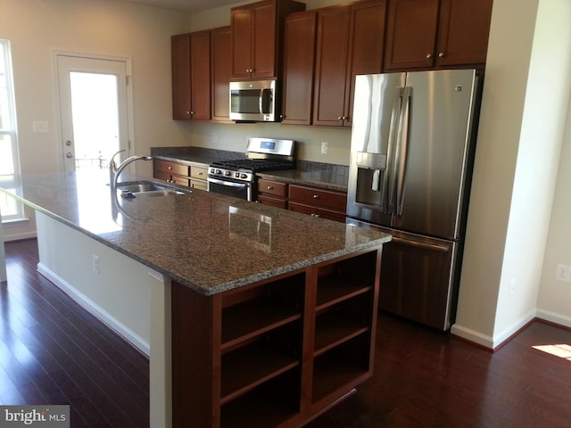 kitchen with sink, dark wood-type flooring, stainless steel appliances, a center island with sink, and dark stone counters