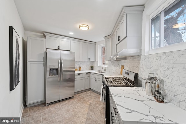 kitchen with light stone countertops, under cabinet range hood, a sink, appliances with stainless steel finishes, and decorative backsplash