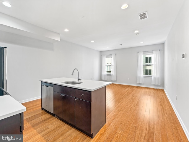 kitchen with an island with sink, sink, stainless steel dishwasher, dark brown cabinetry, and light hardwood / wood-style flooring