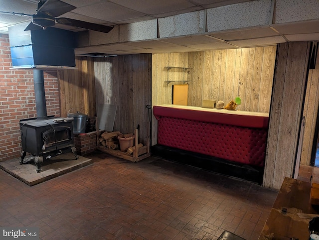 basement featuring ceiling fan, a wood stove, a paneled ceiling, and wooden walls