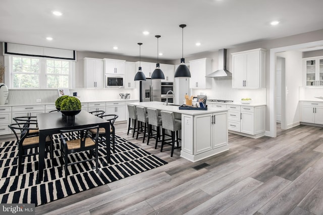 kitchen featuring wall chimney exhaust hood, white cabinetry, hanging light fixtures, a center island with sink, and appliances with stainless steel finishes