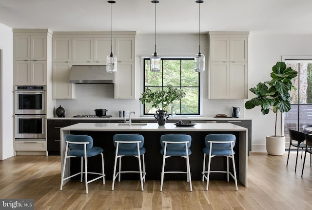 kitchen featuring light wood-style floors, stainless steel double oven, an island with sink, gas cooktop, and under cabinet range hood