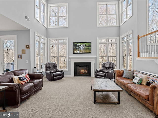 carpeted living area featuring a towering ceiling, visible vents, a wealth of natural light, and a glass covered fireplace