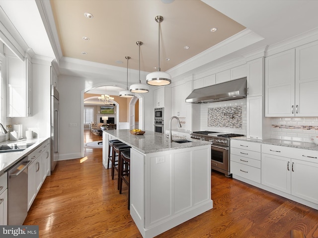 kitchen featuring sink, high end appliances, a raised ceiling, a center island with sink, and wall chimney range hood