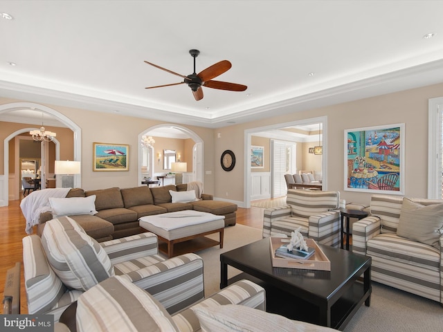 living room with a raised ceiling, ceiling fan with notable chandelier, and light wood-type flooring